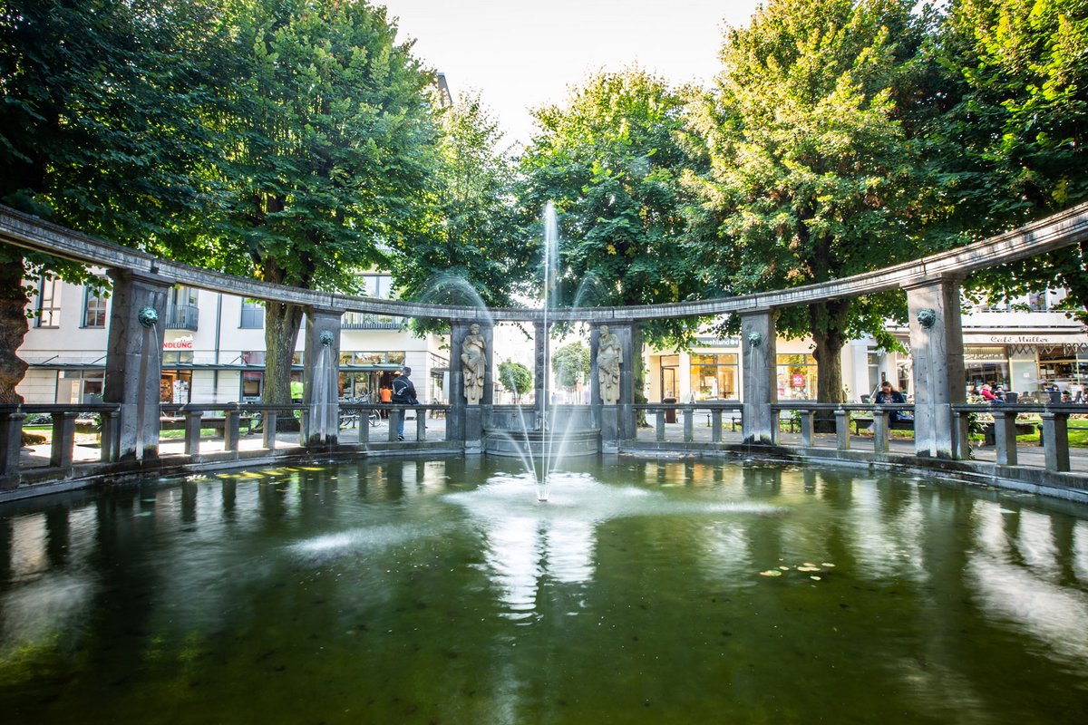 Brunnenanlage in Bad Nauheim mit Wasserfontäne, steinernen Figuren und Arkaden, umgeben von Bäumen und Geschäften.